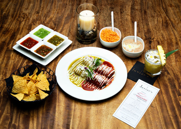 Top-down view of Mexican enchiladas drizzled with red and green sauces and crema on a white plate, plated with tortilla chips, four salsas, rice, refried beans, a pineapple-garnished cocktail and a candle on a rustic wooden table.
