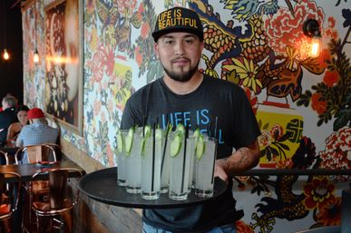 Server wearing a "Life Is Beautiful" cap carrying a tray of cucumber-garnished cocktails in a colorful mural-adorned casual restaurant interior.