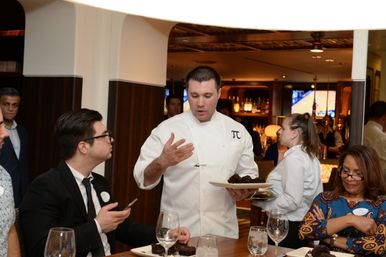 Chef in a white jacket with a π emblem presenting chocolate desserts to seated diners at an upscale restaurant table with wine glasses.