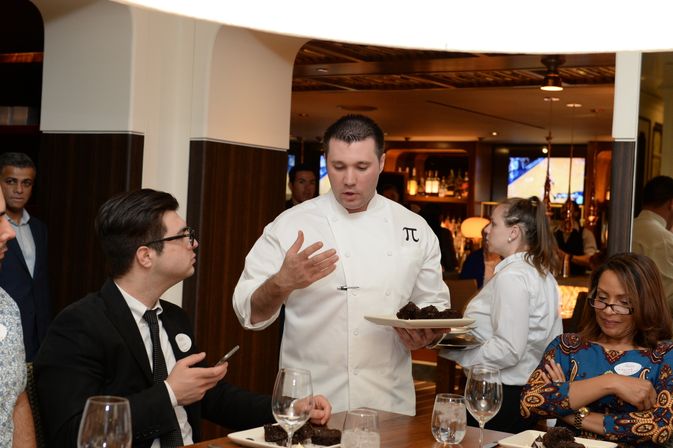 Chef in a white jacket with a π emblem presenting chocolate desserts to seated diners at an upscale restaurant table with wine glasses.