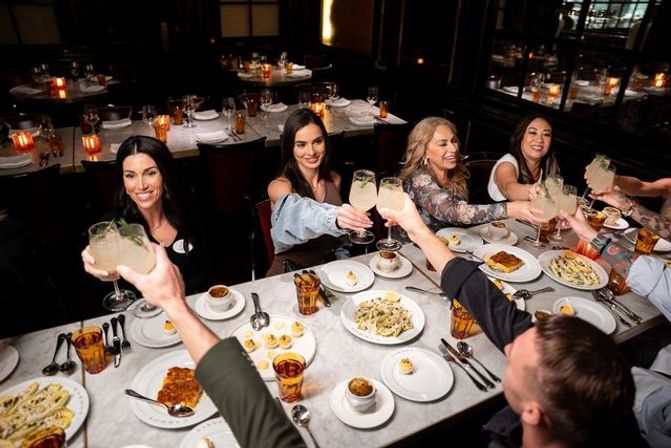 Group of friends toasting cocktails over a marble table with pasta, appetizers and candlelit place settings in a dim, upscale restaurant — lively dinner celebration.