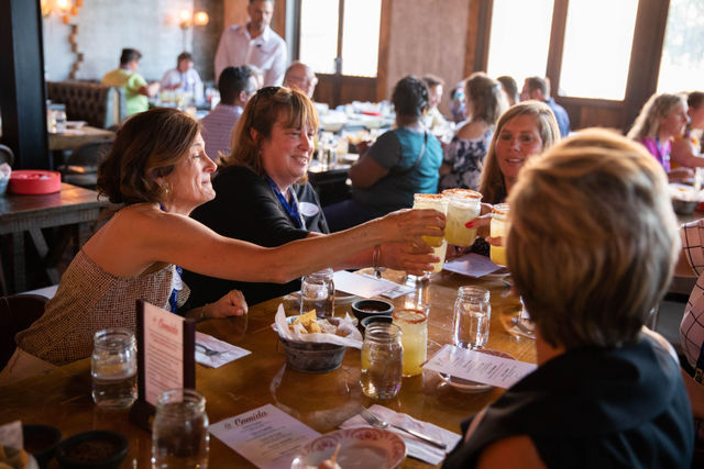 Friends clinking margarita glasses at a busy casual restaurant table with chips and salsa, mason-jar drinks, menus, and warm window light.
