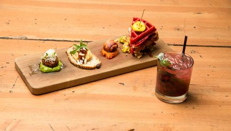 Colorful gourmet appetizer board with four bite-sized tapas (meatball on green puree, crostini, bacon-wrapped bite, mini red waffle sandwich) beside a mint berry cocktail in a glass on a rustic wooden table