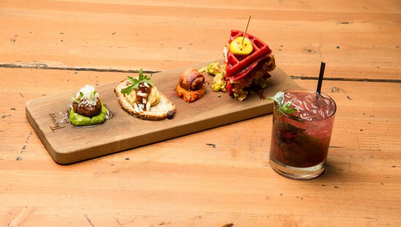 Colorful gourmet appetizer board with four bite-sized tapas (meatball on green puree, crostini, bacon-wrapped bite, mini red waffle sandwich) beside a mint berry cocktail in a glass on a rustic wooden table