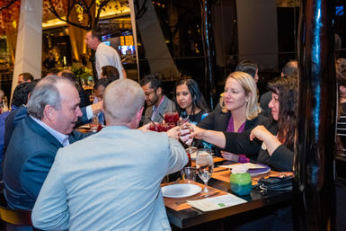Group of professionals toasting with wine and cocktails around a restaurant table during a lively networking dinner, with plates, glasses and a waiter in the background.