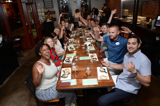 Large group of smiling adults enjoying a lively group dinner at a long wooden table in a cozy restaurant, waving and sharing plates of appetizers and drinks.