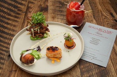 Gourmet appetizer sampler on a white plate — bacon‑wrapped bite, seared seafood cube with orange sauce, crispy croquette and stacked potato chips topped with microgreens, plus a strawberry cocktail and menu card on a rustic wood table.