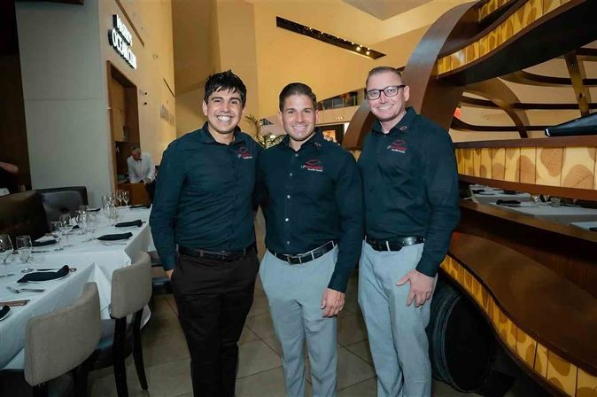 Three smiling restaurant staff in black shirts and gray pants posing by set dining tables in an upscale modern restaurant with a curved wood divider.