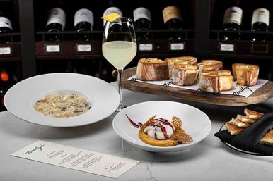Fine-dining spread: creamy mushroom pasta, a scoop of ice cream dessert with berry drizzle, toasted baguette crostini on a wooden board, and a lemon-garnished white wine glass set before a wine-rack backdrop.