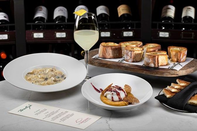 Fine-dining spread: creamy mushroom pasta, a scoop of ice cream dessert with berry drizzle, toasted baguette crostini on a wooden board, and a lemon-garnished white wine glass set before a wine-rack backdrop.