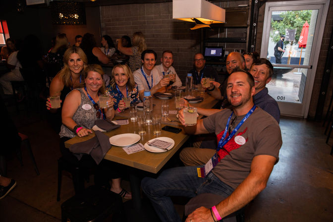 Smiling group of adults wearing conference lanyards enjoying cocktails at a long table inside a dimly lit urban restaurant during a networking happy hour.