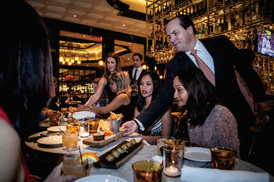Waiter in a suit serving shared appetizers to a group at an upscale restaurant bar — marble table with cocktails, small plates, fries and warm ambient lighting during a lively group dinner.