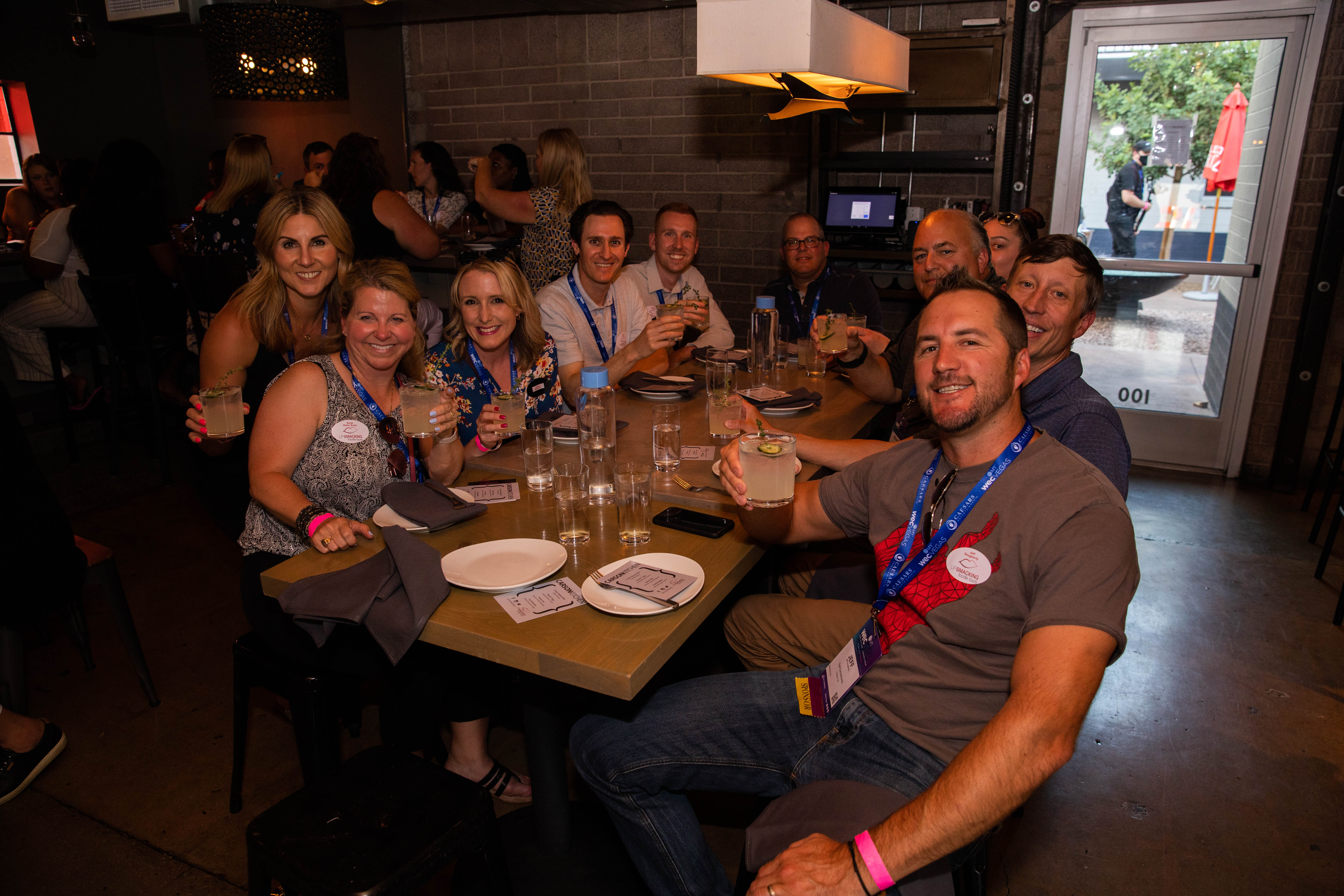 Smiling group of adults with event lanyards clinking cocktails around a wooden table in a casual indoor bar or restaurant with exposed brick and a glass door to an outdoor patio.