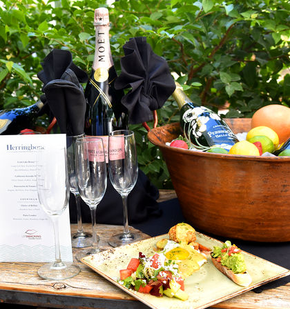 Outdoor garden brunch: chilled champagne bottles in an ice-filled copper bowl, three flutes, a menu card, and a plate of eggs, avocado toast and fresh salad.