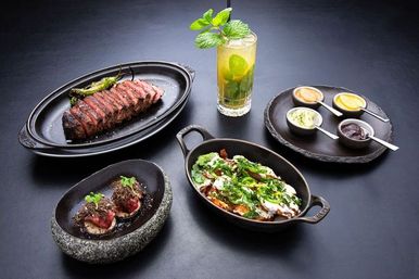 Gourmet restaurant spread on a dark table: sliced medium‑rare steak with charred peppers on a black platter, a cast‑iron skillet of herb‑topped roasted vegetables with creamy sauce, two bite‑sized tartlets in a stone bowl, four dipping sauces, and a tall mint cocktail.