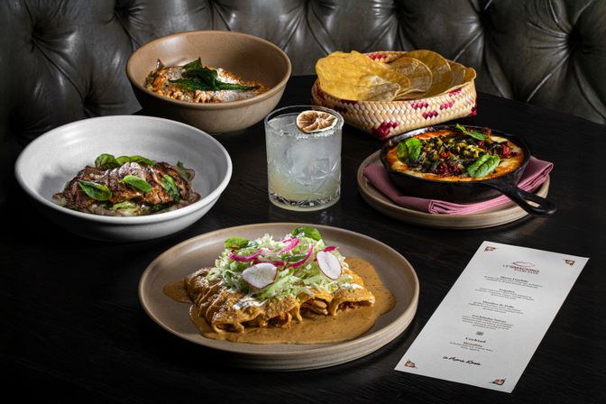 Upscale Mexican restaurant table spread with mole-covered enchiladas topped with shredded lettuce, radish and crema in the foreground, grilled steak bowl, creamy pasta, skillet of melted cheese and chiles, basket of tortilla chips, and a citrus cocktail beside a paper menu.