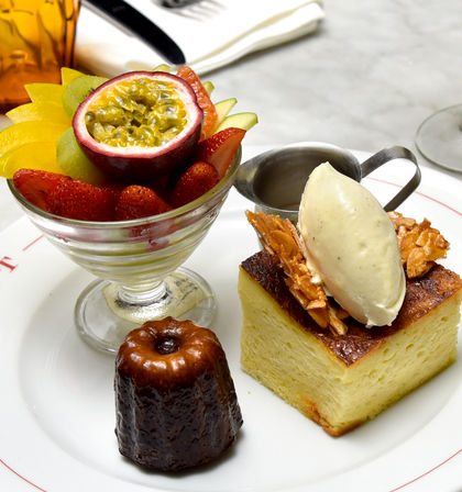 Assorted dessert plate with a glass fruit cup topped with halved passionfruit, strawberries, kiwi and mango, a caramelized canelé, and a square brioche pudding crowned with vanilla ice cream and almond brittle plus a small sauce pitcher on a white plate.
