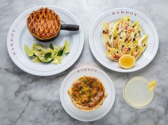Top-down view of three bistro-style dishes on a marble table: a mini skillet with a golden lattice-topped pot pie and lime garnish, endive leaves filled with seafood and lemon, a cheesy baked quiche, and a lemon-twist cocktail.