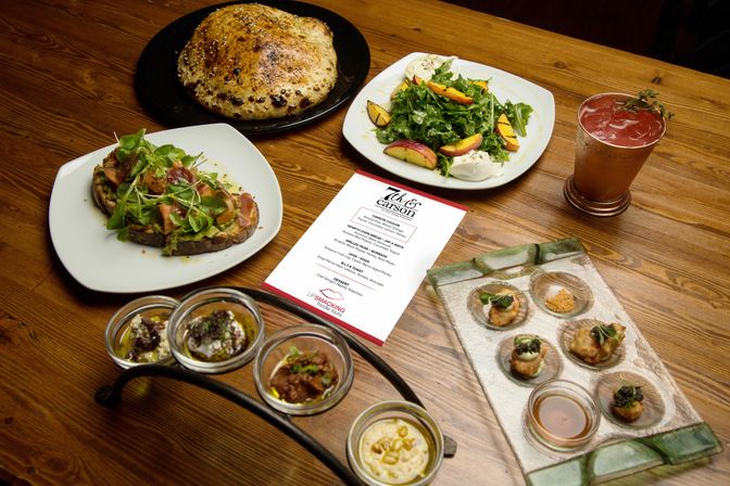 Gourmet restaurant spread on a wooden table: blistered artisan bread, peach-arugula salad with burrata, avocado toast topped with greens, small tasting jars and crostini bites, plus a cocktail and menu card.