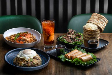 Colorful Thai-inspired spread on a wooden table: fried rice topped with a sunny-side egg, stir-fried vegetables, crispy spring rolls with dipping sauce, marinated grilled beef on lettuce, woven sticky rice basket, and a tall glass of iced tea swirling with milk.