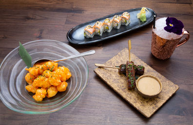 Asian-fusion appetizer spread on a wooden table: spicy popcorn shrimp in a glass bowl, salmon sushi roll with wasabi and ginger on a black platter, grilled beef skewers with dipping sauce on a stone slab, and a copper mug cocktail topped with crushed ice and a purple edible flower.