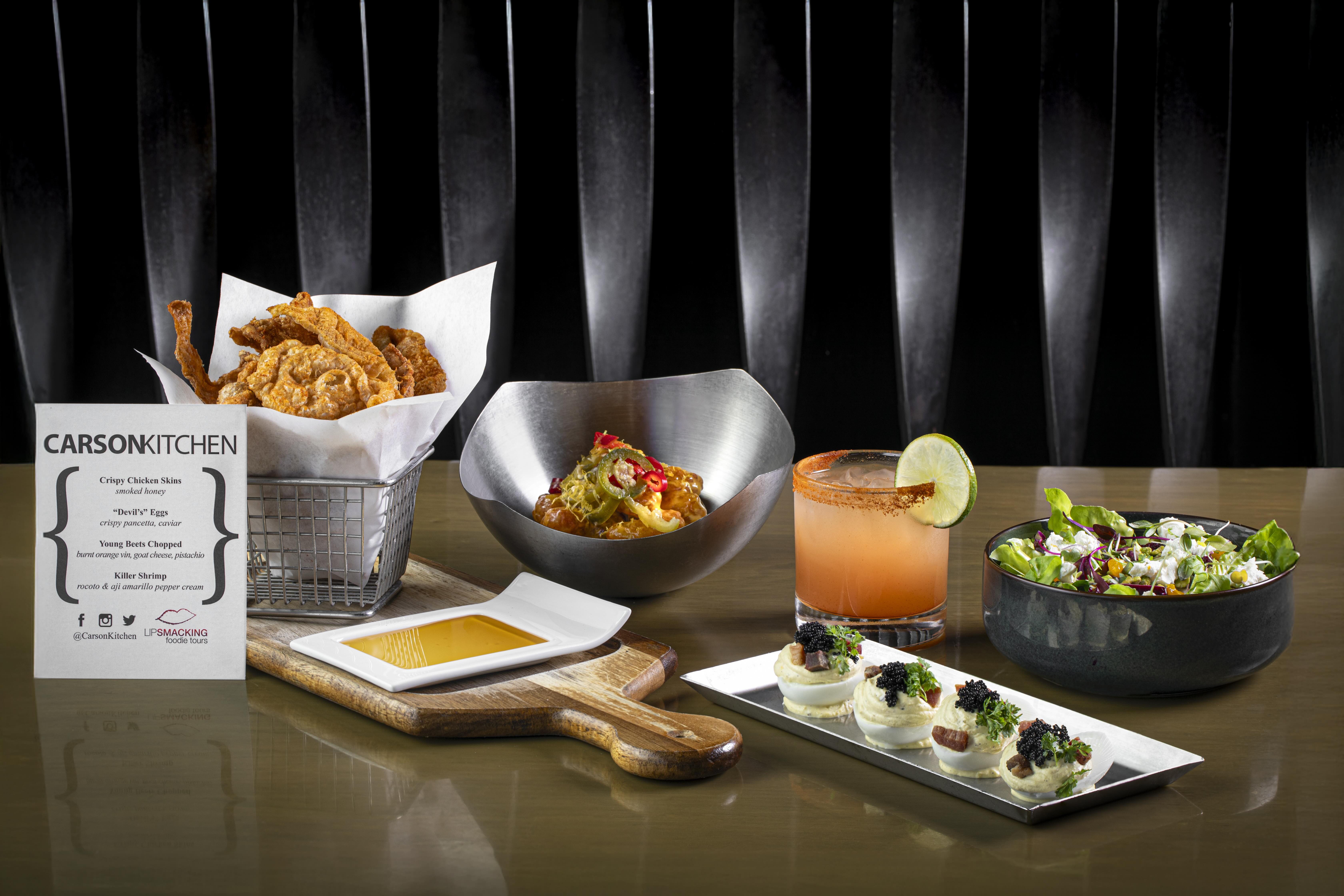 Gourmet restaurant appetizer spread on a glossy table: basket of crispy chicken skins, metal bowl of seasoned bites with peppers, rimmed citrus cocktail with lime, fresh mixed green salad, and deviled eggs topped with caviar and herbs.