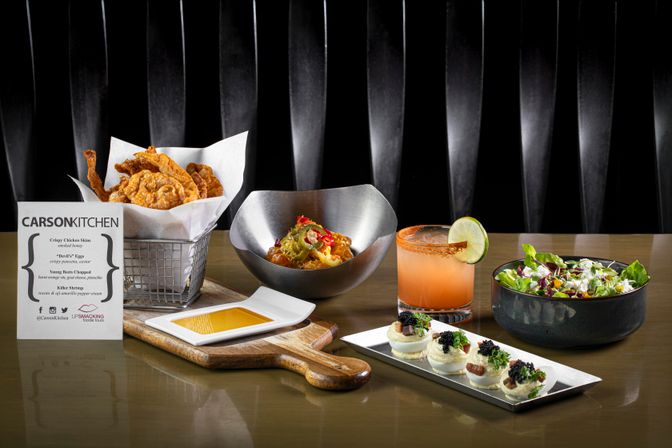 Gourmet restaurant appetizer spread on a glossy table: basket of crispy chicken skins, metal bowl of seasoned bites with peppers, rimmed citrus cocktail with lime, fresh mixed green salad, and deviled eggs topped with caviar and herbs.