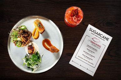 Overhead shot of a gourmet small-plates tasting course on a white plate—seared scallop, mixed greens, crispy garnish and sauce dots—paired with a red craft cocktail and a printed menu on a dark wood restaurant table.
