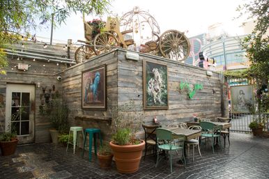 Quirky urban outdoor courtyard cafe with weathered wooden walls, a vintage horse-drawn carriage display on the roof, framed paintings, potted plants, string lights, and mismatched bistro tables and chairs.