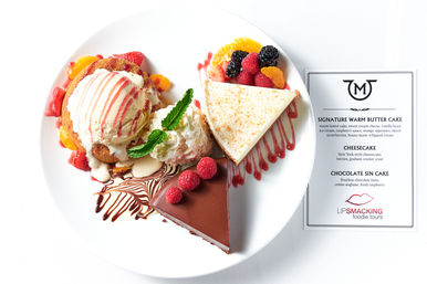 Overhead shot of a dessert plate: vanilla ice cream on warm cake with whipped cream and fruit, a slice of cheesecake with mixed berries and orange segments, and a glossy chocolate slice topped with raspberries and mint, with a dessert menu card beside the plate.