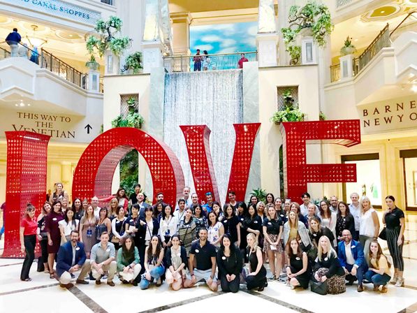 Smiling group poses before oversized red LOVE letters sculpture in an ornate hotel atrium with a cascading waterfall and balconies