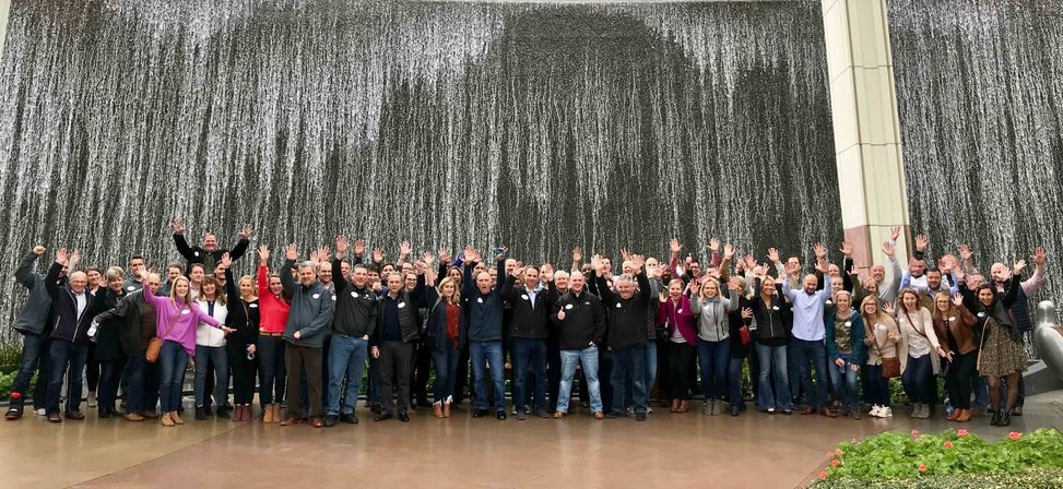 Cheerful large group smiling and waving for a panoramic photo in front of a dramatic cascading waterwall fountain at an urban plaza on an overcast day