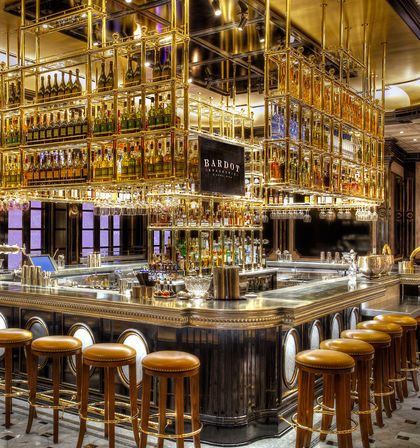 Glamorous luxury hotel bar interior with gold-plated hanging bottle racks, mirrored shelving, marble countertop, and round leather bar stools