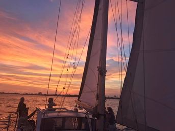 Sunset sail with silhouettes on a sailboat, tall sails and rigging framing vibrant pink-orange clouds over calm harbor waters and a distant coastal skyline.