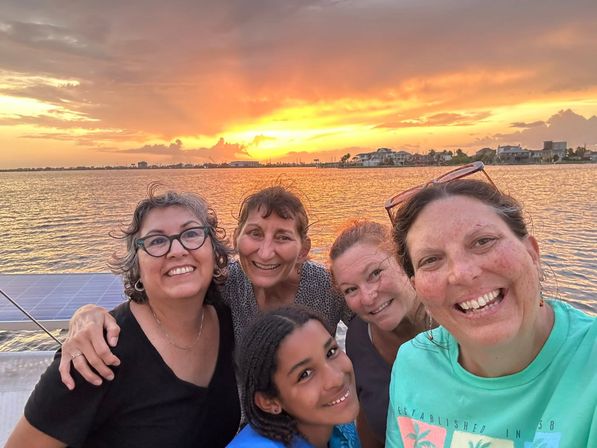 Boat selfie of five smiling people against a golden coastal sunset over a calm bay with waterfront homes on the horizon.