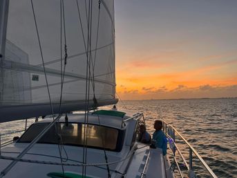 Sunset sail with two people on a sailboat deck, raised sails and rigging silhouetted against an orange coastal sunset over calm open water.