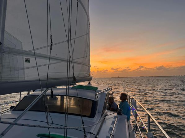 Sunset sail with two people on a sailboat deck, raised sails and rigging silhouetted against an orange coastal sunset over calm open water.