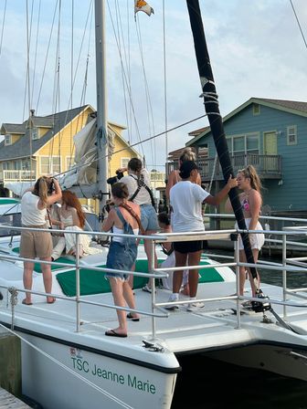 Group of friends on a white catamaran at a coastal marina with photographers shooting and colorful beach houses lining the dock