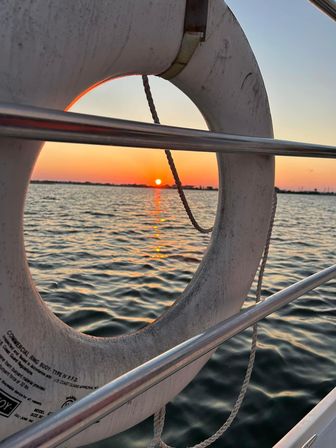 Orange sunset over calm coastal water reflected in ripples, playfully framed like a porthole through a white lifebuoy mounted on a boat’s stainless-steel railing.