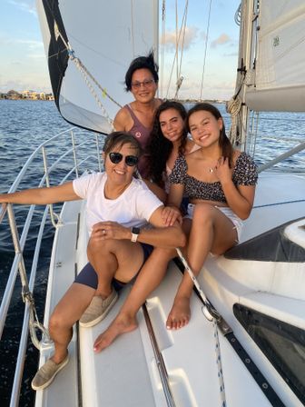 Four smiling people lounging on a sailboat deck near a coastal shoreline at golden hour, sails up and calm water in the background.