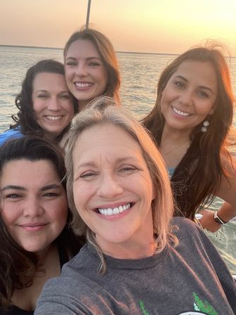 Five smiling women taking a selfie on a boat at sunset, warm golden light reflecting on calm water