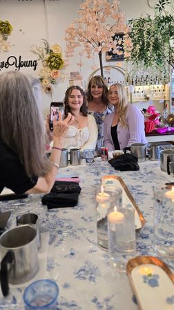 Three women smiling for a group photo inside a boutique floral-themed studio with a faux cherry blossom tree, shelves of bottles, and a candlelit table with metal pitchers and blue floral tablecloth.