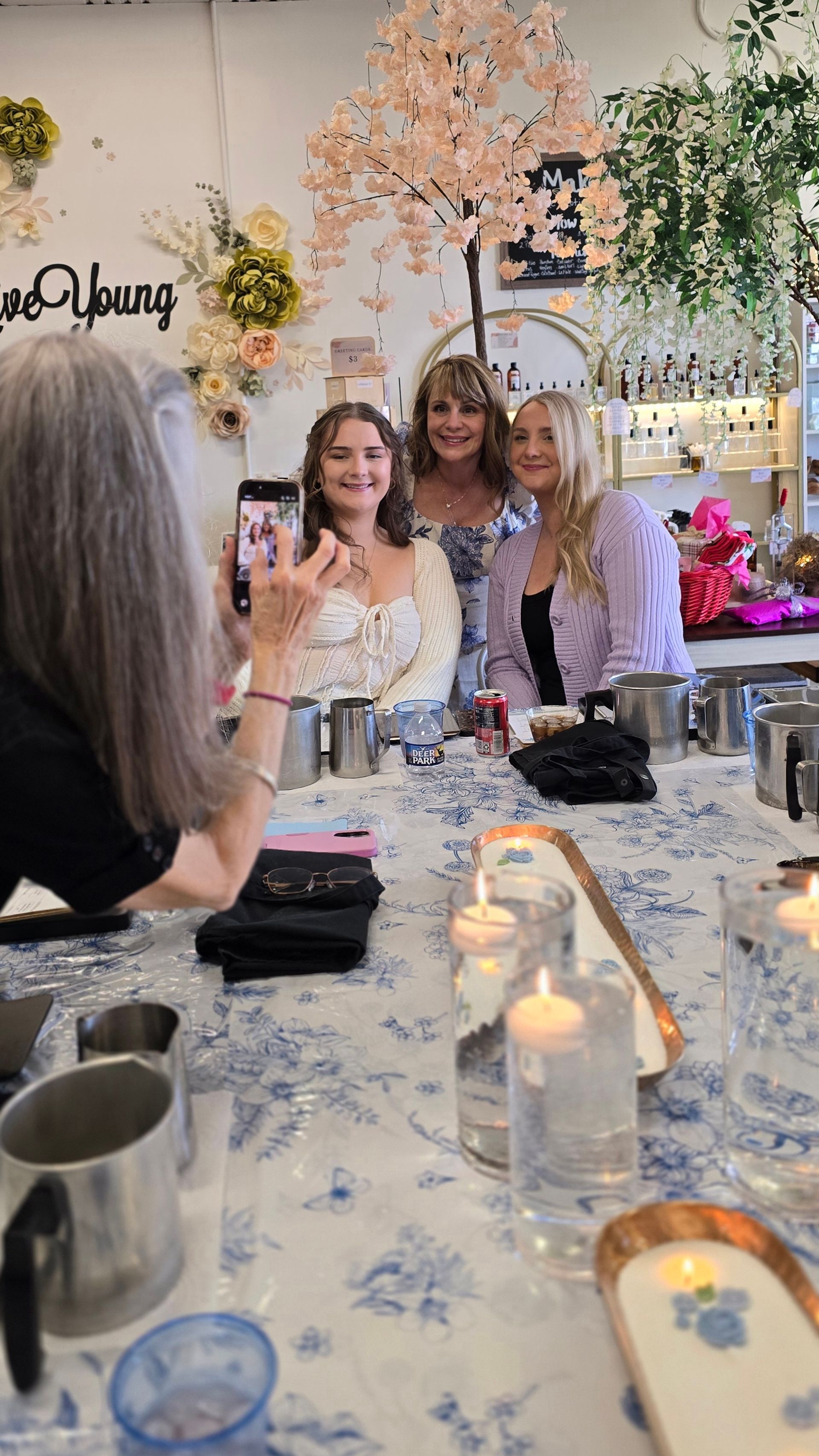 Three women smiling for a group photo inside a boutique floral-themed studio with a faux cherry blossom tree, shelves of bottles, and a candlelit table with metal pitchers and blue floral tablecloth.
