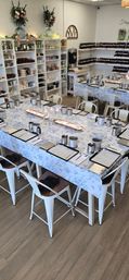 Bright DIY candle-making workshop setup in a cozy craft studio — long tables with blue floral tablecloths, stainless pouring pitchers, digital scales, clipboards, glassware and lit candles, with shelves of jars and fragrance bottles in the background.