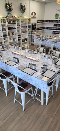 Bright DIY candle-making workshop setup in a cozy craft studio — long tables with blue floral tablecloths, stainless pouring pitchers, digital scales, clipboards, glassware and lit candles, with shelves of jars and fragrance bottles in the background.