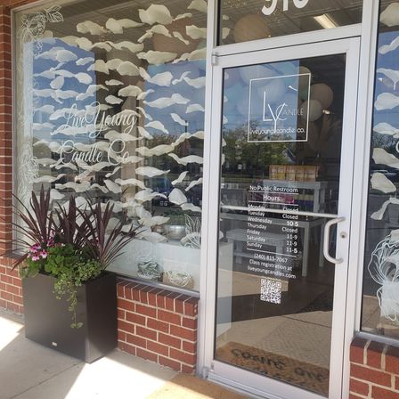 Brick-and-glass storefront with decorative white leaf window decals, glass door displaying hours and a QR code, black planter with purple flowers, and candles and pastel balloons visible inside.