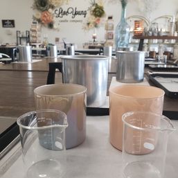 Candle-making workshop setup with metal pouring pots, two 100 ml glass beakers and pastel candle vessels on trays at a communal wooden table in a bright studio