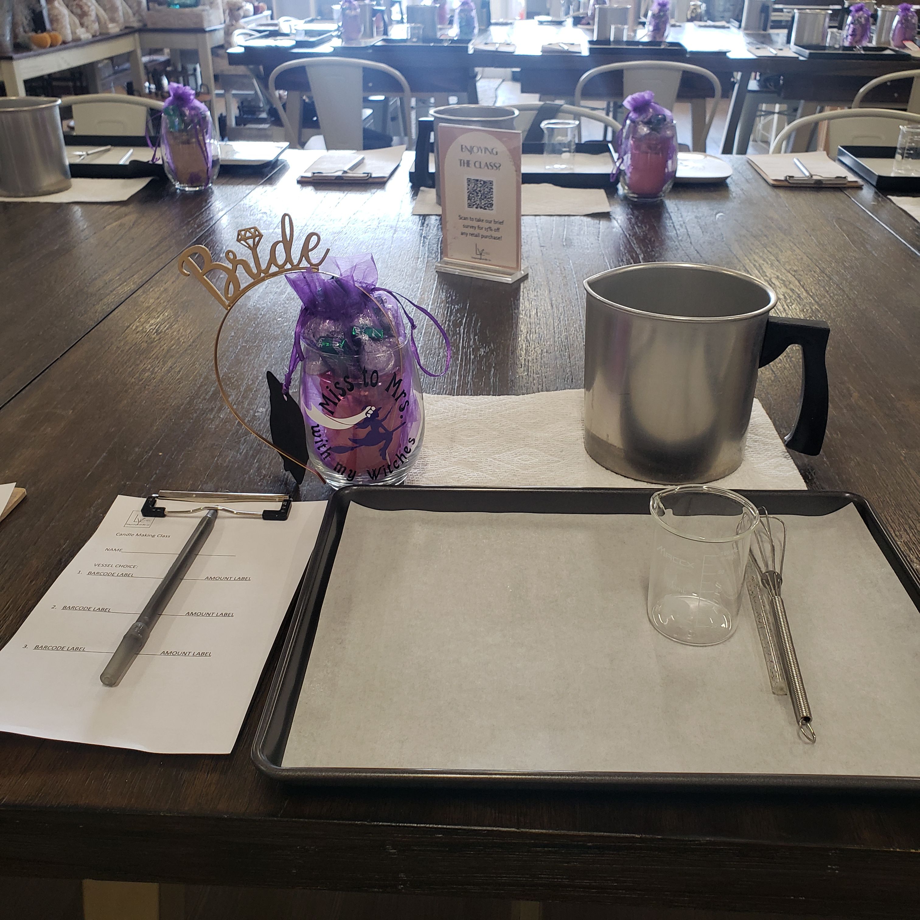 Candle-making workshop table setup in a craft studio with a metal melting pot, glass measuring beaker and whisk on a parchment-lined tray, clipboard and pen, and a purple ‘Bride’ favor bag.