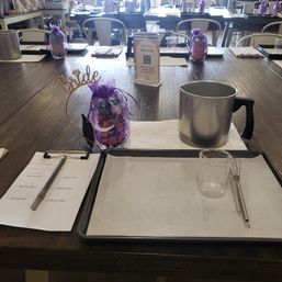 Candle-making workshop table setup in a craft studio with a metal melting pot, glass measuring beaker and whisk on a parchment-lined tray, clipboard and pen, and a purple ‘Bride’ favor bag.