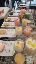 Artisan handmade candles in colorful glass jars with floral wax toppers and bamboo lids arranged on a metal shelving rack with printed labels — craft studio display.